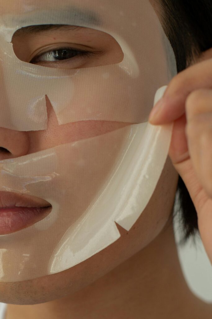 Crop unrecognizable Asian male model looking at camera while taking off moisturizing facial mask during skincare routine in light studio